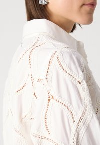 Chemise blanche avec des détails en dentelle intricats, présentant un col pointu et des coutures décoratives. Tissu lisse et léger.