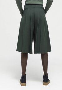 Donkergroene, middellange shorts van een zachte stof. Kenmerken zijn plooien, een wijd beenontwerp en een zachte tailleband. Gecombineerd met donkergouden enkelboots.