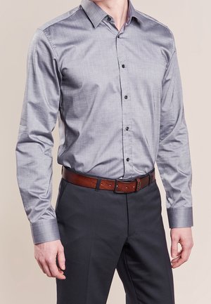 Homme portant une chemise grise cintrée à manches longues boutonnée, un pantalon foncé et une ceinture en cuir marron, debout devant un fond uni.