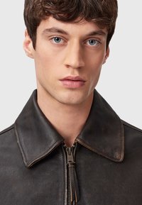 Giacca di pelle marrone scuro con un ampio colletto, cerniera visibile e superficie testurizzata. Presenta dettagli in pelle e un design strutturato.