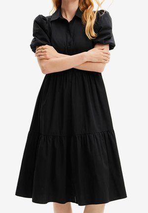 Femme portant une robe midi noire avec des manches courtes bouffantes et un col, les bras croisés sur la poitrine.