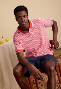 Rood gemêleerd poloshirt met blauwe accenten, voorzien van een klassieke kraag. Draagt het met donkere shorts. Model zit op een houten stoel.