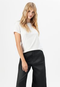 Femme blonde aux cheveux ondulés portant une chemise blanche à manches courtes et un pantalon noir taille haute, sur fond blanc.