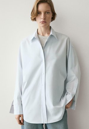 Jeune personne aux cheveux courts portant une chemise oversize bleu clair boutonnée par-dessus un haut gris, debout contre un mur blanc uni.