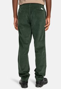 Green corduroy pants with a straight cut, featuring two back pockets and a small logo tag, paired with black sneakers.