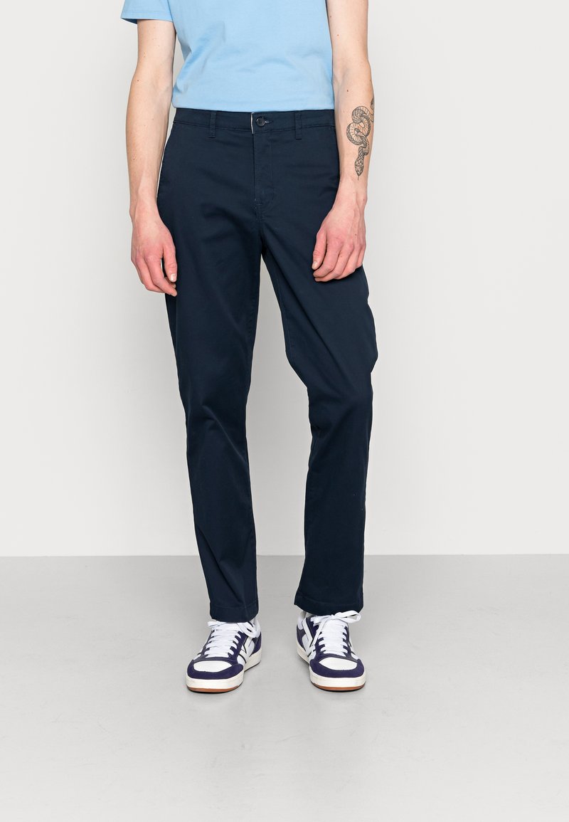 Marineblaue Chinos aus Baumwolle mit geradem Bein, vorderen Taschen und Knopfverschluss, kombiniert mit weißen und lila Sneakern.