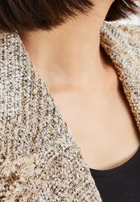 Texturerad ljusbrun cardigan med gyllene och svarta trådar, med en lös passform och öppen krage, bärs över en svart topp.