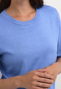 Persona che indossa un maglione a maniche corte di un azzurro chiaro, con le mani gentilmente incrociate in vita, tagliato a livello del collo e a metà torace su sfondo bianco.