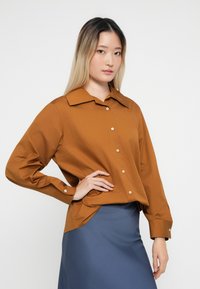 Ung kvinde med langt ombre hår iført en brun skjorte med knapper og en blå nederdel, poserer med den ene hånd på hoften mod en hvid baggrund.