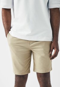 Pantalones cortos de algodón color caqui con corte recto y dobladillo doblado, que presentan bolsillos laterales y una textura suave, combinados con una camiseta gris claro.