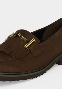 Mocassin en daim marron avec boucle métallique dorée et détail à franges sur le dessus, doté d'une semelle épaisse en caoutchouc foncé.