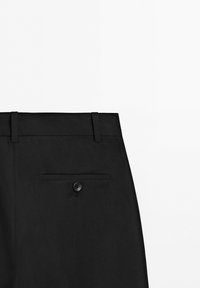 Vista posteriore di pantaloni eleganti neri che mostrano la cintura, due passanti e una singola tasca posteriore con chiusura a bottone nero.