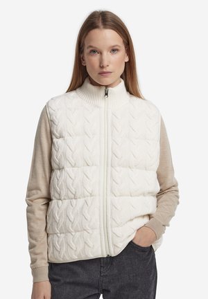 Frau mit langen Haaren trägt eine weiße, gesteppte Strickweste mit Reißverschluss über beigefarbenen, langärmligen Oberteilen, eine Hand in der dunklen Denim-Tasche, vor weißem Hintergrund.