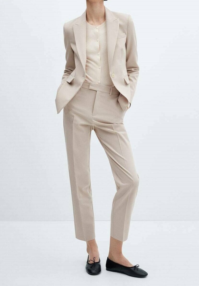 Beżowy garnitur z jednorzędową marynarką, slim fit spodniami i jasnobeżowym topem. Czarne baleriny z kokardą. Gładka faktura.