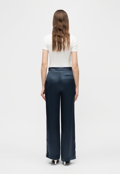 Γυναίκα με μακριά κυματιστά μαλλιά, φορώντας λευκό κορμάκι με κοντά μανίκια, μπλε-σκούρες φαρδιές σατέν παντελόνες και λευκές γόβες, στέκεται πλάτη σε απλό φόντο.