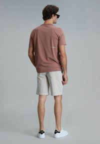 Korte mouwen bruine t-shirt met een ronde hals, voorzien van een klein logo op de achterkant, gecombineerd met lichtbeige shorts en witte sneakers.