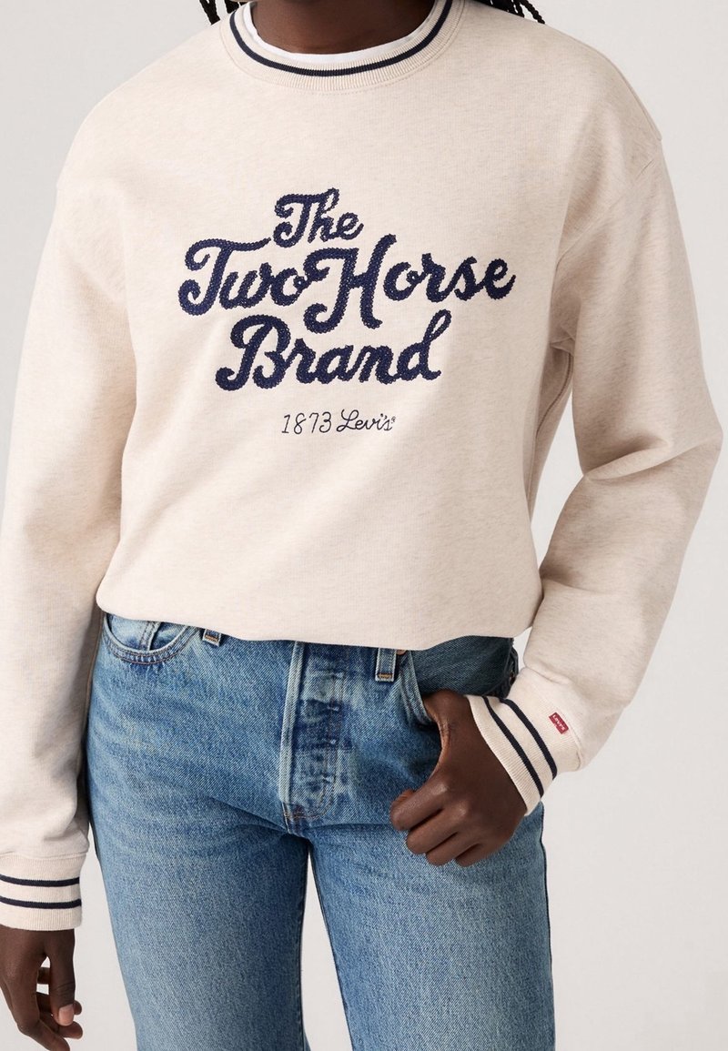 Personne portant un sweat beige avec un texte bleu marine "The Two Horse Brand 1873 Levi's" et un jean bleu, main dans la poche.