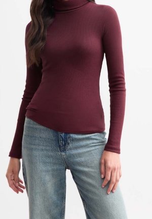 Femme portant un col roulé bordeaux moulant à manches longues et un jean taille haute bleu clair sur un fond uni.