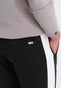 Zwarte broek met een witte zijsstrik, voorzien van een klein logo-label op de zak. De stof lijkt glad en heeft een aansluitend ontwerp.