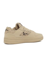 Beige színű sneaker, sima szintetikus anyagból, párnázott nyelvvel, díszes, domború mintázattal és fekete logóval az oldalon. Lapos gumitalp.