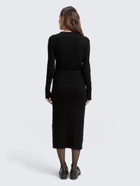 Robe noire à manches longues avec un corsage cintré et une longueur mi-mollet. Texture lisse et fente sur le côté, assortie à des collants opaques et des chaussures noires.