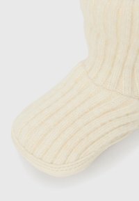 Botte tricotée de couleur crème avec une texture côtelée, un bout arrondi et un matériau doux avec des coutures visibles le long de la semelle.