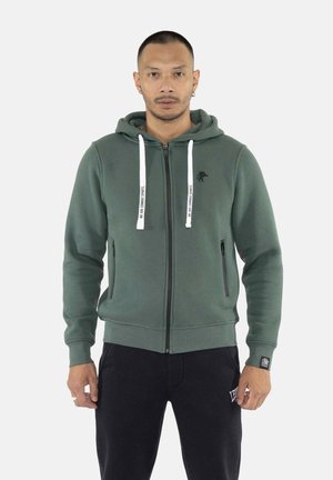 Hombre con sudadera verde con cremallera y cordones blancos y pantalones negros, de pie frente a un fondo claro y liso.
