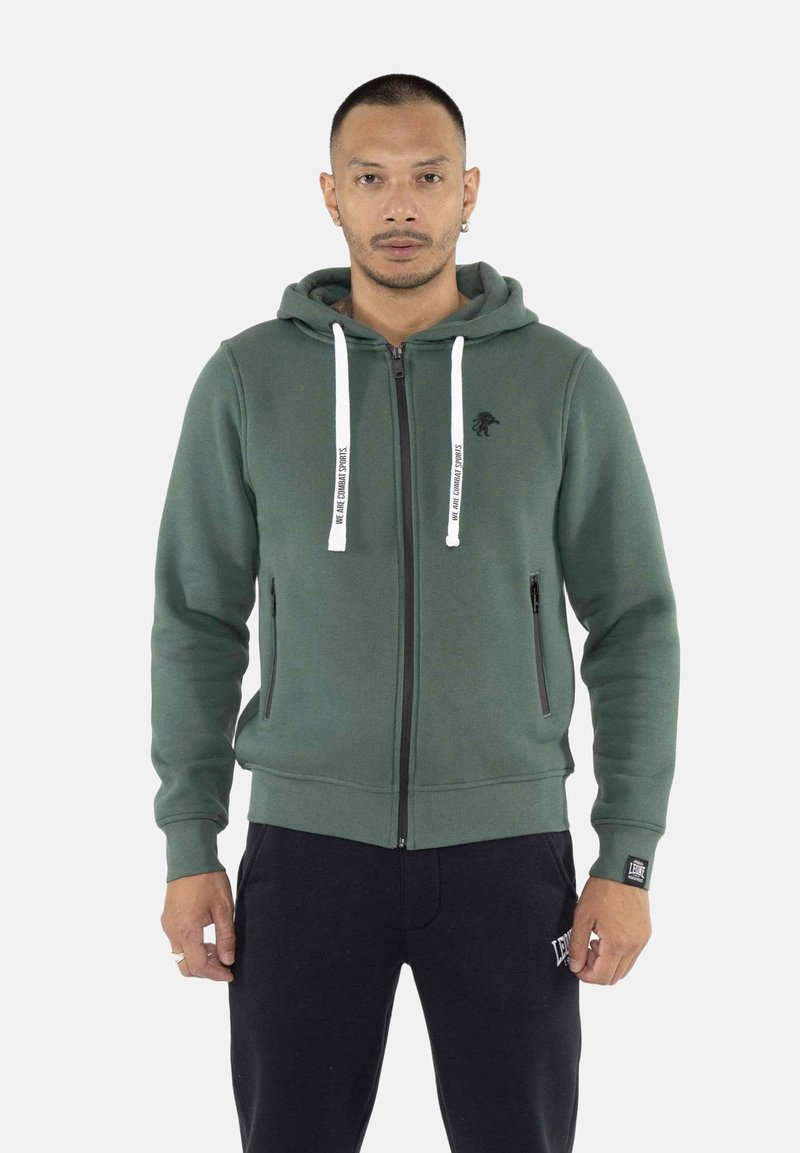Homme portant un sweat à capuche zippé vert avec des cordons blancs et un pantalon noir, debout devant un fond clair uni.