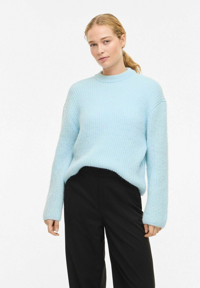 Hele sinine soonikkoes kampsun kõrge kaelusega ja pikkade varrukatega, valmistatud pehmest, tekstuuriga materjalist, kombineeritud mustade pükstega.