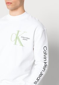 Sudadera blanca con un logo verde de "CK" y el texto "Calvin Klein Jeans" impresos en el pecho y en la manga izquierda. Textura suave.