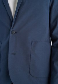 Κοντινό πλάνο ενός μπλε μπλεζάκι με ένα κουμπί κουμπωμένο και μια μεγάλη επιδιορθωτική τσέπη στη δεξιά πλευρά πάνω από ένα λευκό πουκάμισο.