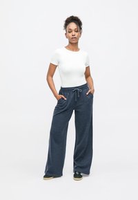 Γυναίκα στέκεται με τα χέρια στις τσέπες, φοράει λευκό εφαρμοστό τοπ με κοντά μανίκια, φαρδιά σκούρα γκρι παντελόνια με κορδόνι και μαύρα αθλητικά παπούτσια με ταμπά σόλες.