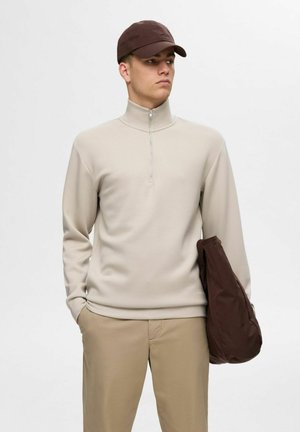 Beige halv-zip sweatshirt med lange ærmer, glat tekstur. Lys brun cap og brun taske holdt i venstre hånd. Slim-fit beige bukser.