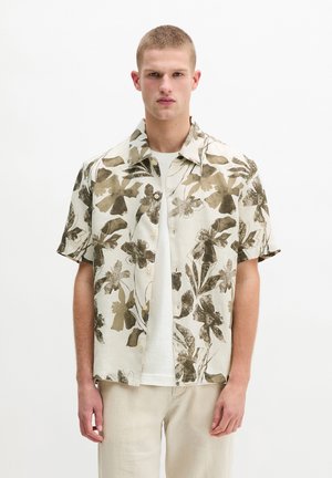 Młody mężczyzna z krótkimi włosami, ubrany w beżową koszulę w kwiaty z krótkim rękawem i guzikami, na białą koszulkę i jasne spodnie, stojący na białym tle.