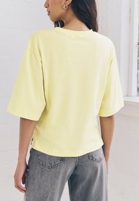 Locker sitzendes T-Shirt mit kurzen Ärmeln aus einem hellgelben Stoff, mit rundem Halsausschnitt und leicht gebogenem Saum, kombiniert mit grauen Jeans aus Denim.