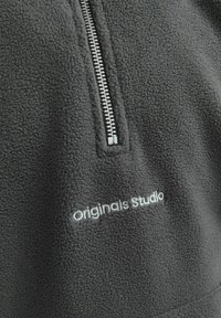 Pulover iz flisa v temno sivi barvi s pol zadrgo. Ima mehko teksturo in vezeno "Originals studio" logo v svetlo sivi barvi.