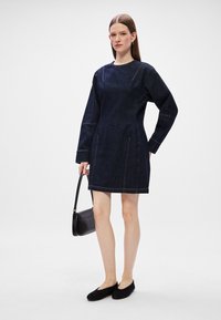 Σκούρο τζιν φόρεμα με μακριά μανίκια, στρογγυλή λαιμόκοψη και ορατές λεπτομέρειες ραφής. Φορεμένο με μαύρες φλατ παπούτσια και μια μικρή τσάντα ώμου.