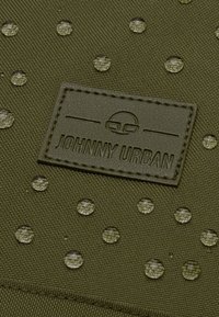 Zöld textúrált szövet, amely kiemelkedő kör alakú mintákkal és egy téglalap alakú bőr folttal rendelkezik, amelyen "JOHNNY URBAN" van gravírozva.