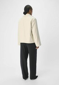 Manteau en mélange de laine de couleur claire avec une coupe décontractée, doté d'un col cranté et de longues manches, associé à un pantalon sombre et des chaussures pointues.