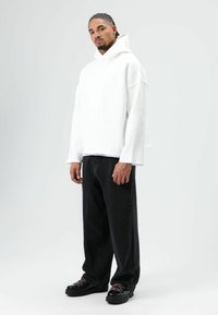 Sweat à capuche blanc oversize avec manches larges et tombantes, associé à un jean noir à jambes larges et à des chaussures noires à lacets avec des accents roses.