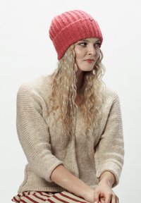 Różowy dzianinowy kapelusz z prążkowaną teksturą i subtelnym niebieskim akcentem. Noszony z beżowym, grubym swetrem. Czerwono-białe pasy na dole.