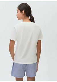 White cotton t-shirt with short sleeves, crew neckline, smooth texture, paired with blue and white striped shorts, shown from the back.