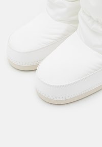 Chaussures rembourrées blanches avec une texture lisse, un bout rond et une semelle extérieure crème. Présente des détails couturés le long des bords.