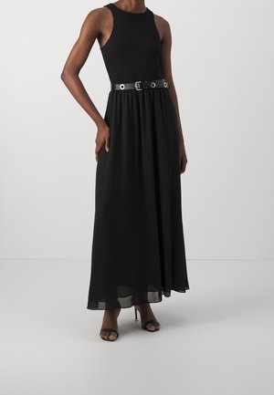 Femme portant une robe longue noire sans manches avec un haut texturé, une taille ceinturée avec des œillets métalliques, et des sandales à talons noires ouvertes.
