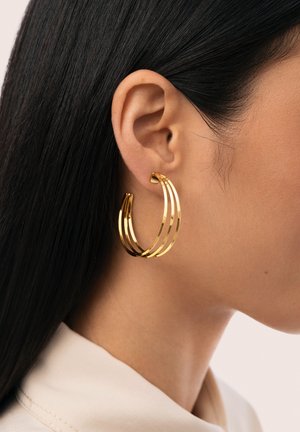 Primer plano de la oreja de una persona con cabello largo y negro, que lleva un gran aro de oro compuesto por tres bandas conectadas.