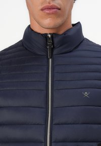 Homme portant une veste matelassée bleu marine avec une fermeture éclair argentée et un petit logo de canne croisée et chapeau sur le côté gauche de la poitrine.