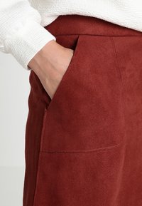 Jupe en daim bordeaux avec une texture lisse, dotée de poches latérales et d'une taille haute, partiellement recouverte par un vêtement texturé blanc.