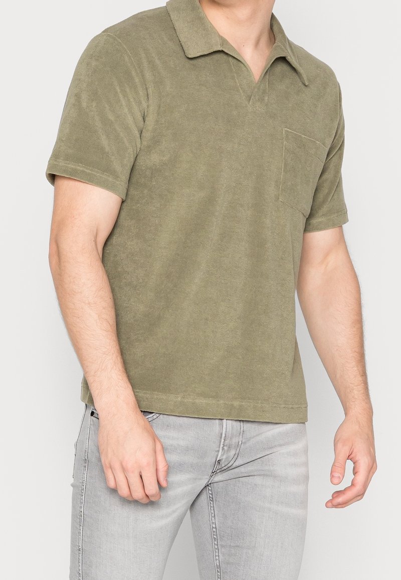 Homme portant un polo vert olive à manches courtes avec une poche poitrine et un jean gris clair, debout devant un fond uni.