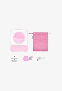 Dispositif de nettoyage facial en silicone rose avec surface texturée, emballage, câble USB et sac à cordon assorti présentés sur un fond blanc.