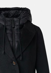 Cappotto nero con cappuccio staccabile imbottito, dotato di chiusura con zip, cordini e un bottone decorativo nella parte anteriore. Tessuti testurizzati in tutto il capo.